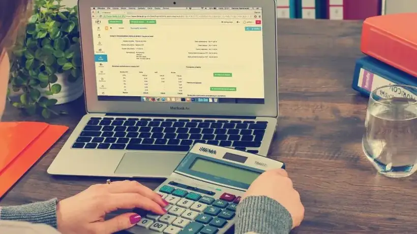 A person using a calculator beside a laptop showing a spreadsheet